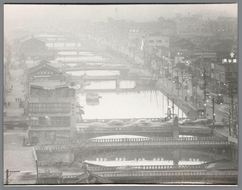 The image depicts a bustling urban scene with multiple layers of infrastructure. At the foreground, there is an elevated train system spanning horizontally across the frame, indicative of rapid transit in densely populated cities like Osaka.

Below this layer, we see rows of traditional Japanese buildings, characterized by their wooden structures and thatched roofs. These edifices are tightly packed together along a canal or river which reflects faint light from nearby sources, creating soft illumination on its surface. 

In the middle ground, there's another train line running parallel to an elevated road where cars can be seen moving in both directions. The architecture of these buildings appears more modern compared to those directly beside the waterway.

The background features high-rise apartment blocks and commercial structures with lights that hint at a vibrant nightlife or active business hours for the cityscape depicted here. These towering buildings, lit up against an overcast sky suggest it might be evening time in this urban setting of Osaka. The entire scene is captured from a vantage point above street level providing a panoramic view of various modes and layers of transportation within this metropolitan area.

There's Japanese writing visible on the right-hand side of the image which could provide additional context or location markers, possibly indicating street names, company logos, or even advertisement [...]
