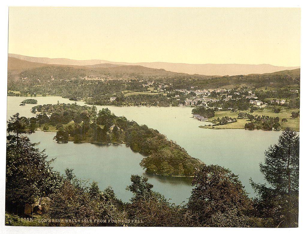 The image depicts a picturesque, aerial view of a serene lake landscape. The water appears tranquil and reflects the surrounding greenery, with various patches of land jutting out into it. A network of tree-covered islands or peninsulas is scattered throughout the scene, suggesting dense woodland areas. Lush trees and shrubs border the shoreline, indicating abundant vegetation typical of temperate regions.

Several clusters of buildings are visible on some parts of the shorelines, which could be houses, cottages, or smaller structures such as boathouses, indicative of human habitation in a rural setting. The architecture suggests traditional British design with pitched roofs and possibly stone construction.

The backdrop is composed of gentle rolling hills that merge into a faint outline of distant mountains under a pale sky transitioning from yellow to blue at the horizon line, which could imply either dawn or dusk lighting conditions. The overall color palette consists of muted greens, blues, browns, and yellows, giving it an aged appearance.

The image quality is reminiscent of early 20th-century postcards, with possible signs of damage such as creasing along one side, yellowing edges indicating age, and a faint watermark or label at the bottom left corner. These suggest that this might be an old photograph from around the late 1800s to early 1900s.

A handwritten insc [...]