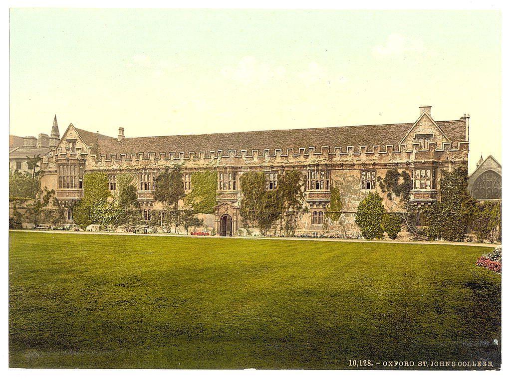 The image depicts a large, historic building with architectural features reminiscent of medieval or Renaissance styles. The structure is characterized by its extensive use of stone masonry and ornate detailing around the windows, doors, and roofline. Ivy clings to parts of the facade, adding a touch of greenery against the earthy tones of the brickwork. In front of the building lies an expansive lawn with vividly colored grass, suggesting meticulous maintenance. The sky is clear, indicating fair weather conditions at the time the photograph was taken. A watermark or label in one corner provides attribution to Oxford St John's College and includes a numerical sequence likely used for cataloging this image within its collection.