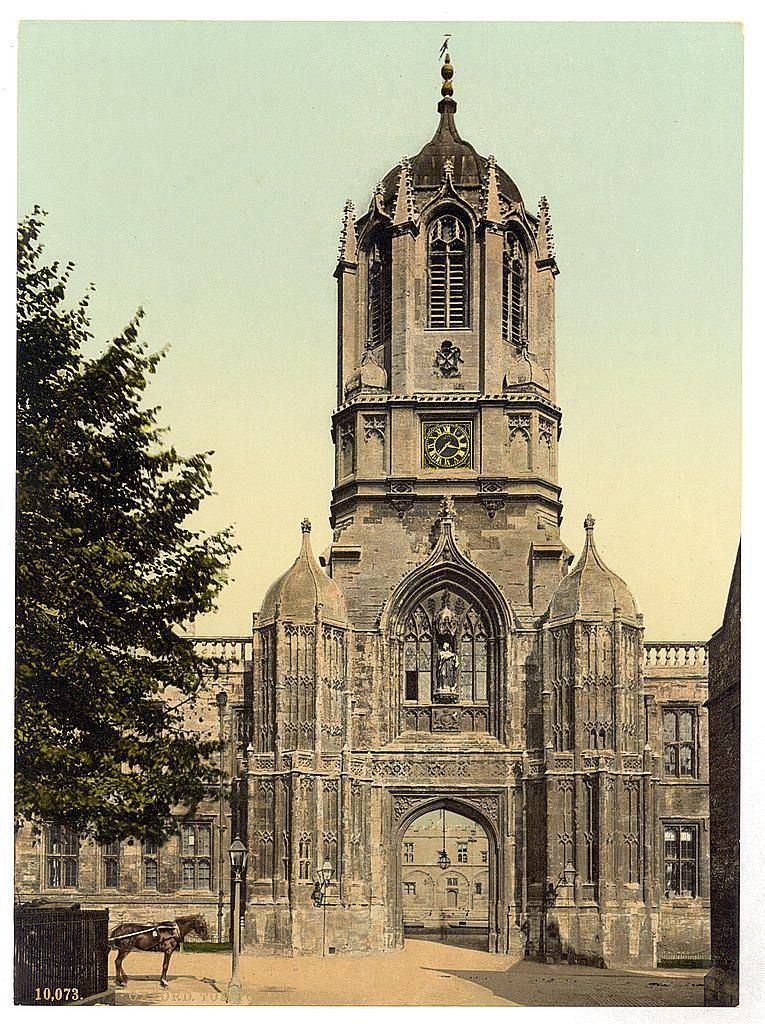 The image depicts a large, historic stone building with Gothic architectural features. Prominently standing in the center is an ornate clock tower that extends upwards into the sky, capped by a weather vane or finial. The facade of the structure showcases pointed arches and intricate stonework, indicative of its medieval design. A series of tall windows punctuate the walls at different levels, some with stained glass designs visible through their transparency.

The building has multiple openings: an arched entranceway in the middle section leading into a larger courtyard or inner space; another opening to the left side facing directly towards us; and smaller doorways on other parts. It appears aged, as evidenced by the patina of weathering that gives it a sense of history and longevity.

In front of this imposing structure stands a horse-drawn cart with two horses attached at either end, suggesting an earlier time when such conveyances were common for transportation or delivery purposes within urban settings like Oxford in England. The surrounding environment includes mature trees to the left side which frames part of the scene and provides some shade on what seems to be a sunny day.

The ground is paved, indicative of city infrastructure rather than rural countryside, emphasizing its likely role as an important public building such as a university or town hall during tha [...]