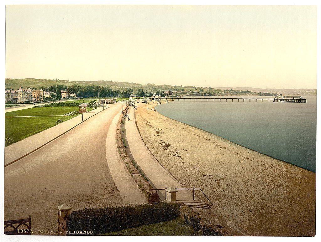 The image depicts a serene coastal scene, likely from an early 20th-century postcard. It shows Paignton Sands in England with a view of the beach extending towards calm waters and beyond that to a long pier or jetty on the right side. The shoreline curves gently as it approaches what appears to be a promenade leading into town. On this path, there are small figures of people enjoying their day out.

In the foreground, we see well-maintained grass lawns with flower beds and benches alongside paths that guide visitors along the beachfront. There's an ornate street lamp post on the left side close to the road. The background reveals rolling green hills, indicating a hilly terrain beyond the town area. Buildings can be seen in the distance near these hills.

The overall atmosphere of the image is tranquil and picturesque, capturing a typical leisurely day at this seaside resort during its heyday as a holiday destination.
