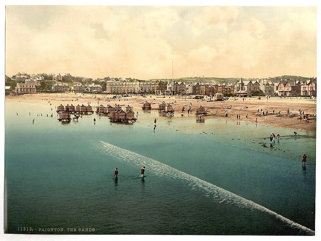The image depicts a scenic view of Paignton Beach in the English town of Paignton, as suggested by both textual annotations and the visual context. The photograph is likely from around 1890 to 1900 based on stylistic elements like sepia tones and period-specific architecture.

It shows a beachside landscape with numerous people scattered across sand and water areas engaging in various leisure activities such as walking, playing, or bathing. In the foreground, two individuals are wading through shallow waters leaving behind waves that create a foamy trail extending towards the shore.

The middle ground displays groups of wooden structures on stilts floating just above the water's surface; these could be bathing huts or similar beachside accommodations typical in seaside resorts during this era. The background is lined with rows of late 19th-century architecture, including large buildings likely used for residential and commercial purposes such as hotels.

The image captures a serene yet lively atmosphere indicative of leisurely activities common at the turn of the century along English coastal towns like Paignton. This particular photograph serves as both an artistic record and social history artifact, showcasing typical beachside scenes from that period in British life.
