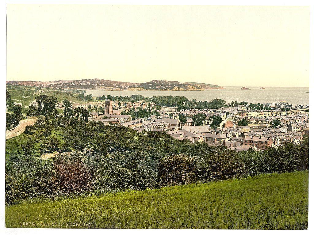 The image is a historical photograph depicting an expansive view of the coastal town situated between Torquay and Paignton in England. It appears to be from around 1890-1900, given its style and coloration.

In the foreground, we see lush greenery with dense vegetation including trees and bushes on one side and a well-maintained grassy area on another. The middle ground showcases an extensive urban landscape of Torquay or Paignton, characterized by numerous closely packed buildings, mainly residential houses but also some larger structures that could be public institutions like churches.

The town's layout is dense with roads winding through the center, and there are a few prominent trees breaking up the monotony. The architecture suggests European style housing from late 19th-century Britain. There’s an absence of modern vehicles or electric infrastructure indicating it may have been taken during a time when these were not widespread.

The background reveals a vast expanse of sea with small islands and possibly ships at some distance, hinting at the town's connection to maritime activities. The sky is slightly hazy, which could be due to atmospheric conditions typical for coastal areas or perhaps a result of photographic processing from that era.

Overall, it’s an image capturing not only geographical features but also offering insights into urban planning and architectu [...]
