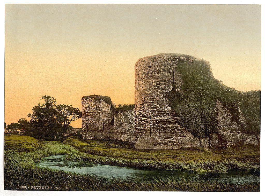 The image depicts a picturesque landscape featuring an old, partially ruinous castle known as Pevensey Castle. The ruins are composed of tall stone walls that have overgrown with ivy and greenery in some areas, suggesting significant aging or abandonment. To the left side of the frame is another imposing structure resembling a tower or keep, which also exhibits signs of age but retains more definition compared to its neighboring remains.

The setting sun casts warm hues across the scene, painting the sky in gradients from yellow near the horizon to softer shades as it ascends towards twilight blue higher up. This lighting condition adds an atmosphere of tranquility and nostalgia to the image.

In front of the castle ruins is a small body of water that appears to be a moat or stream; its surface reflects parts of the surrounding landscape, indicating calm conditions. The foreground is dominated by lush green grass, which contrasts with the brownish tones of the stones. This verdant environment suggests the location could experience mild and temperate climate.

Overall, this image captures not only the architectural remnants but also evokes a sense of history and natural beauty that characterizes such historical sites in England.