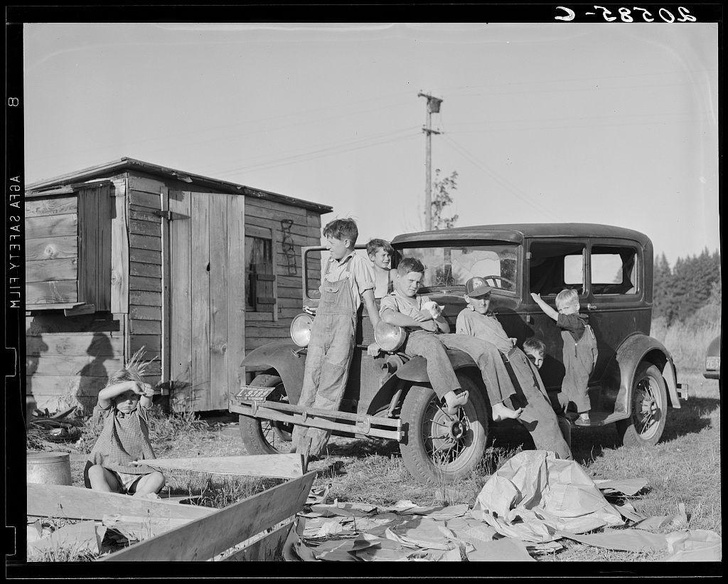 This black and white photograph depicts a group of five young boys standing beside an older model car, with one boy sitting on the ground. The setting appears to be rural or semi-rural, as indicated by the presence of a wooden shed in the background and scattered debris around them. One of the boys is holding what seems to be a bundle of sticks or branches over his head, while others are leaning against various objects nearby.

The car's make and model aren't identifiable from this angle, but it appears well-used with signs of weathering. The overall atmosphere suggests hard labor in agriculture, as hinted by their attire which looks practical for work such as picking beans—hence the title "Bean pickers' children."

There is a young child to the left side sitting on wooden beams or planks, and another one kneeling behind them holding what appears to be pieces of paper. The environment seems quite bare with no trees in close proximity, which might indicate that this photo was taken during harvest time when vegetation has been cleared away.

The photograph carries an emotional weight suggesting a hard life for these children, likely partaking in laborious work alongside their parents or guardians. This aligns with the historical context of migrant farmworkers and rural poverty depicted by Dorothea Lange's work during that period, reflecting societal issues around economic i [...]
