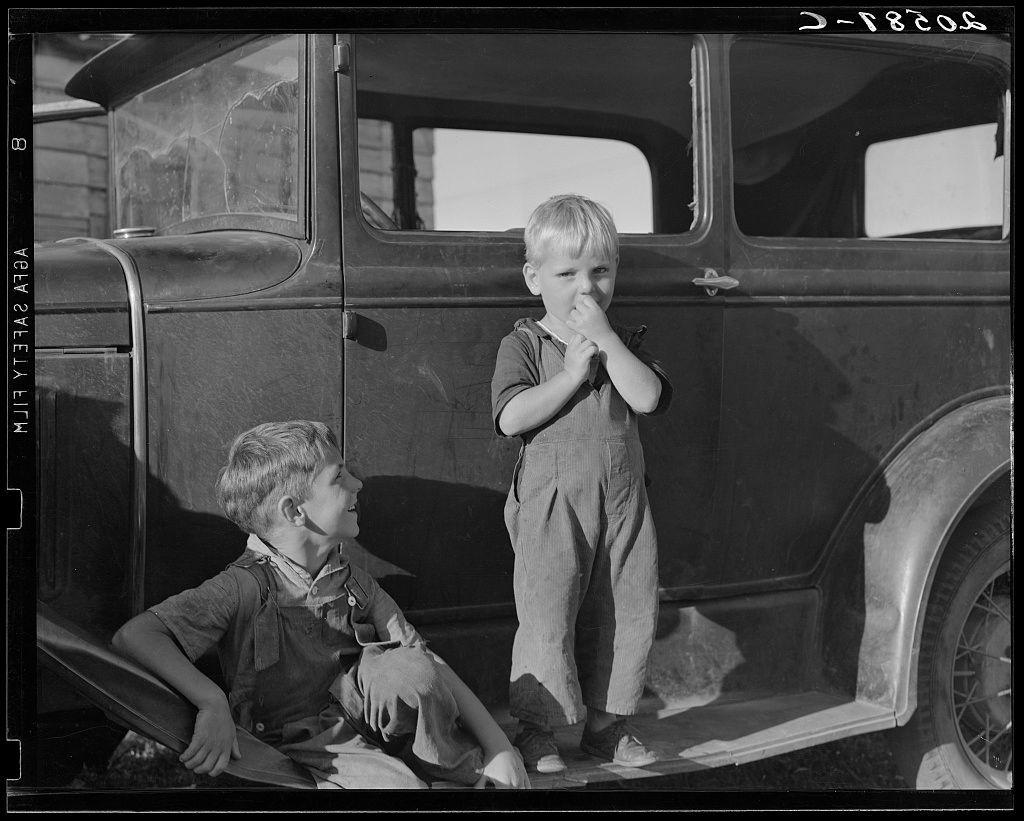 The image is a black and white photograph depicting two young boys next to an old-fashioned car. The setting appears to be outdoors, possibly on the side of a road or in a rural area given the presence of what seems like dust or debris around them.

On the left, one boy is seated inside the vehicle with his legs hanging over the edge. He has short hair and wears rolled-up pants with suspenders, along with a vest that appears to be made of rough fabric. His expression is neutral as he gazes off into the distance.

Standing on the right side near the car's door frame is another boy standing casually in what looks like work clothes or overalls. He has short hair and his attire suggests practicality for outdoor activities, possibly related to farmwork given context clues from other captions mentioned.

The automobile itself appears aged with a rugged design typical of early 20th-century vehicles. It's difficult to determine the make and model due to resolution limitations but it seems like an older sedan type vehicle without any visible branding or significant modern features such as rearview mirrors or windscreen wipers in view at this angle.

Additional information about the image suggests these boys may be associated with a community of agricultural laborers, possibly engaged in picking beans given their attire and setting. This scene likely reflects life during an era whe [...]