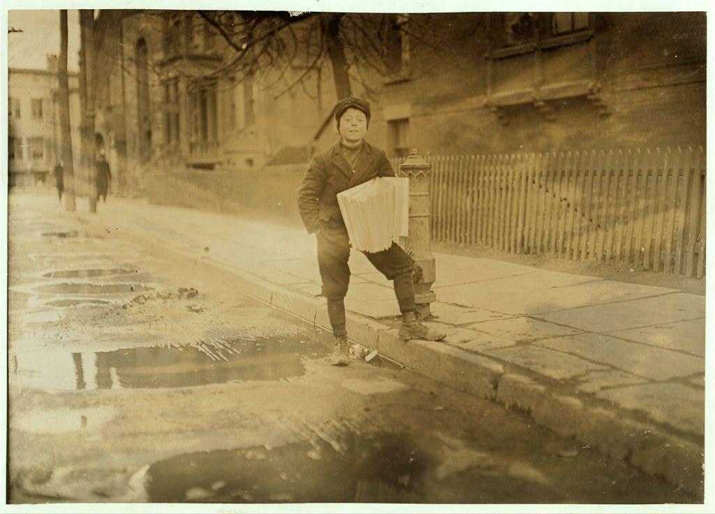The image is a sepia-toned photograph of a young boy standing on the sidewalk in an urban setting. He appears to be around 12 years old and dressed warmly, suggesting it might be cold weather. The boy has a determined expression as he carries what seems to be folded newspapers under his arm while balancing a small box or package with one hand. His feet are clad in heavy boots that appear worn out.
The background shows an older city street lined with bare trees indicating winter season. There's visible snow on the ground, and puddles reflecting light suggest recent thawing of accumulated ice. A fence runs along the side of the building to his right while another person can be seen walking away from him in the distance down the sidewalk.
The image carries historical significance as it is tagged with additional information at 7:00 a.m., suggesting that he sells newspapers before school, after school and on weekends including Saturdays and Sundays. It also indicates an ongoing period of three years since his involvement in this activity. The location mentioned is Bridgeport, Connecticut which gives context to the scene's geographical setting.
The photograph belongs to Lewis Hine whose work often focused on documenting labor conditions particularly during early 20th century America. This image captures a moment from that era providing insight into children’s working lives duri [...]