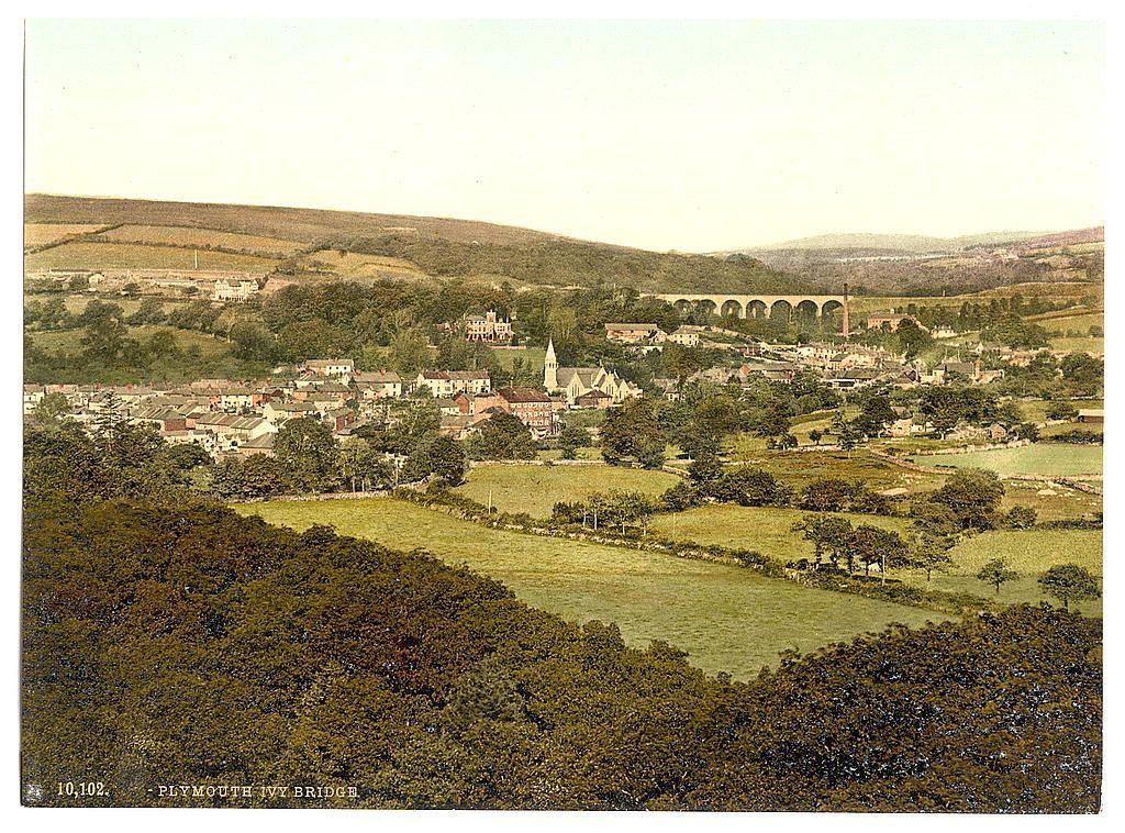 This image depicts a picturesque rural town with diverse architecture, including residential homes, churches, and possibly institutional buildings. A prominent feature is the ivy-covered bridge that spans across what appears to be a river or stream in the foreground. In the background, there's an expansive landscape of gently rolling hills adorned with trees and patches of cultivated land, suggesting agricultural activity. Notably, a railway viaduct can be seen stretching across part of the town, indicating connectivity for transportation during this period.

The overall impression is one of tranquility and historical charm, capturing life in what seems to be late 19th or early 20th-century Plymouth Ivy Bridge, England. The quality of the image suggests it might have been taken using a photographic process like photolithography or photochrom printmaking common during that era.

Further context provided by the reference indicates this view is specific to Plymouth Ivy Bridge in England and possibly part of a larger collection documenting various sights across Britain's Isles between approximately 1890-1900.