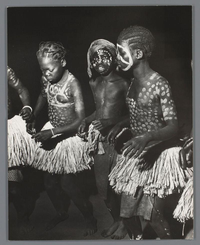 The image captures a group of young children adorned in traditional attire, specifically grass skirts or kava. They are engaged with what appears to be strands of some natural fiber material, possibly preparing for a dance performance known as 'Ganza'. The photograph is monochromatic and exhibits high contrast between the subjects' skin tones against their dark surroundings. Their bodies are marked by white paint in patterns that suggest cultural significance; these markings include dots on various parts of the body such as shoulders, chest, and neck. Notably, one child's face displays a distinctive pattern around the eyes made with black material or paint, creating an intriguing visual contrast to the lighter skin tone.

The children exhibit different expressions: concentration, curiosity, and engagement in their task are evident through their postures and facial expressions. The lighting is dramatic, focusing on the group while leaving much of the surrounding environment in shadow, which draws attention to them as subjects of cultural significance. Their attire and body paint suggest a celebration or ceremony that involves dance and performance elements typical of indigenous traditions.

The image's composition centers around these children, with their grass skirts adding texture and depth to the photograph. There is no indication of modernity beyond this traditional se [...]