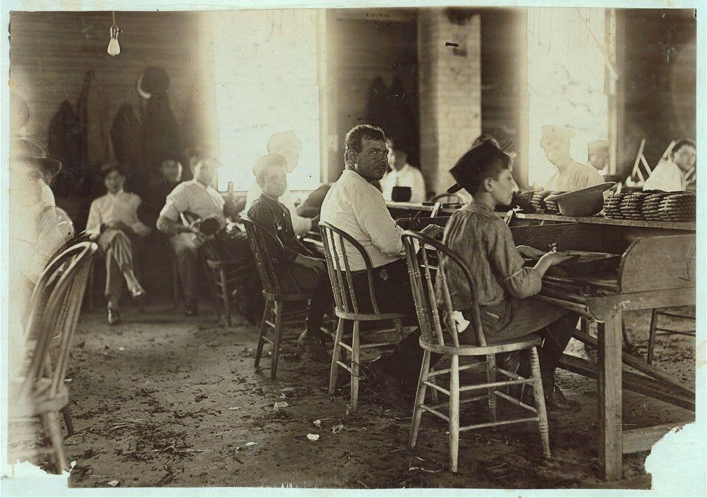 The image is a black and white photograph depicting an indoor scene with several individuals seated around wooden tables. The setting appears to be a workshop or a communal space where people are engaged in various tasks, possibly related to cigar-making given the context provided by the caption.

In the foreground, there's a young boy wearing a wide-brimmed hat sitting at one of the tables, focused on his work with papers spread out before him. He is surrounded by other individuals who seem to be involved in different activities such as reading or working on similar tasks. The people are dressed in casual clothing typical for an early 20th-century environment.

The background shows a row of chairs and more workers seated at their respective tables, suggesting a crowded workspace with limited space between the individuals. There is natural light coming from windows along one side of the room, illuminating parts of it but also casting shadows on other areas. The overall atmosphere appears to be industrious yet humble.

The caption "These boys were surely under 14" indicates that at least some of these young workers are underage and possibly engaged in labor beyond their legal or safe working-age limit for children during the time period implied by the photograph's appearance. This aligns with historical documentation on child labor practices before stricter regulations wer [...]