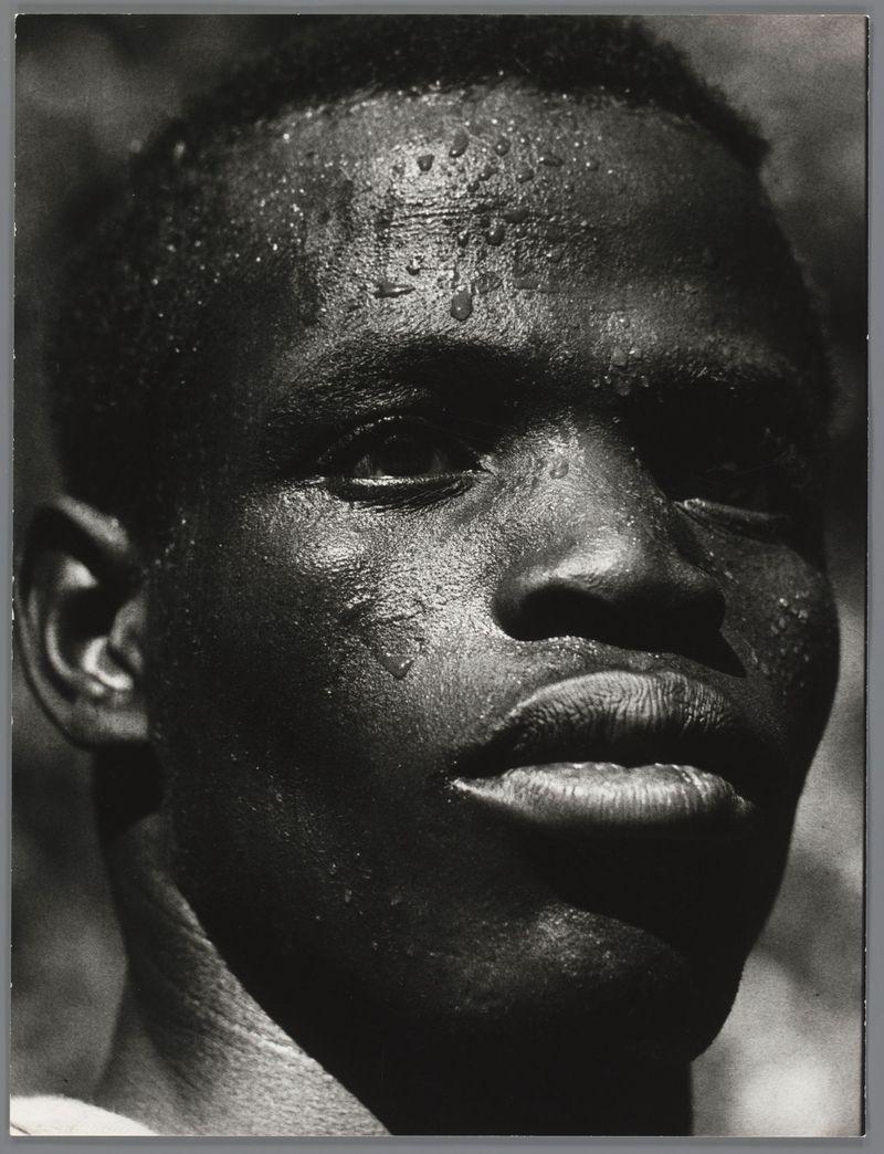 The image is a black and white photograph focusing on the intense gaze of an individual. The person appears to be heavily sweating, with visible droplets on their face that suggest exposure or physical exertion in warm conditions. This effect emphasizes the contours and texture of their skin. There's no distinct background detail due to the close-up nature of the shot; it fades into darkness around the edges, putting full emphasis on the person’s facial features.

The composition captures a moment with deep emotional resonance, as if conveying the fatigue or determination in someone who is possibly engaged in laborious activity, likely related to agriculture given the context provided by another description mentioning 'karavaandrager,' which translates from Afrikaans to "water carrier." The absence of color draws attention to textures and contrasts within the image.