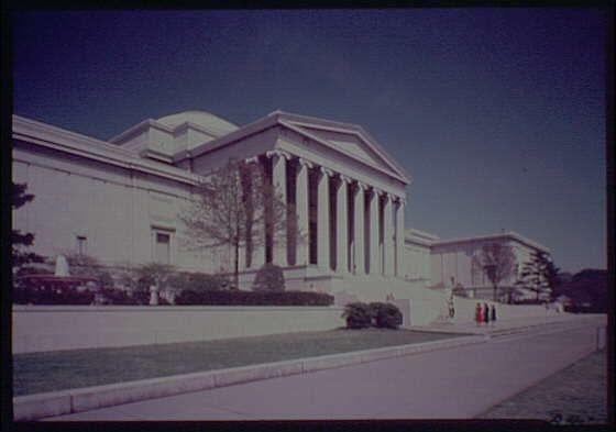 The image depicts the exterior of a large, neoclassical building with prominent Greek Revival architectural features. It has tall columns in front and is situated on an expansive lawn bordered by trees and shrubs. The sky above appears clear blue, suggesting good weather conditions at the time of filming or photography. In the foreground, several individuals can be seen walking along a path leading up to the building's entrance, dressed in what seems like vintage clothing indicative of mid-20th century fashion styles.