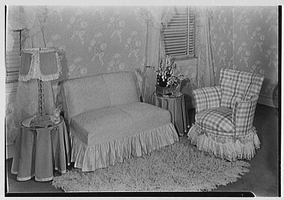 The image depicts a vintage living room setting with classic furnishings and decor from the early 20th century. The main subjects include two upholstered chairs, one featuring a light-colored fabric with a frilled skirt and trimmings, while the other showcases plaid checkered patterned upholstery with similar fringe details at its base.
In between these chairs stands a small wooden table holding an arrangement of flowers in a vase. To the left is another piece of furniture, possibly a stool or side table, adorned with curtains on all sides that create a tent-like appearance around it, and what appears to be a lampshade perched atop it. The entire scene rests upon a patterned carpet extending across the floor.
The background showcases wallpaper with floral patterns, contributing to an overall aesthetic of domestic coziness from past decades. Windows dressed in vertical blinds are partially visible behind these textiles, allowing for natural light diffusion into the room.