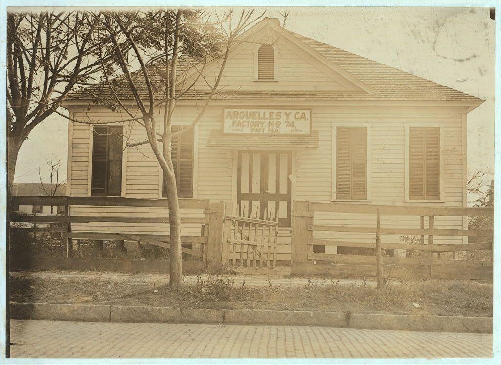 The image depicts a historical building with the sign "Arguelles y Ca., Factory No. 71, Factories Dist. Fla." on its side facade, indicating it is associated with Arguelles and Company in Florida. The structure appears to be an old factory or workshop from the late 19th century or early 20th-century period, featuring a wooden construction with pitched roof, double-hung windows, and shuttered doors.

The photograph has a sepia-toned aesthetic typical of older photographs, suggesting that it is quite old itself. A fence runs along the front yard, separating the building from a paved sidewalk in what appears to be an urban or semi-urban setting. There are bare trees visible near the structure, which may indicate winter time when the photo was taken.

The image does not show any people or active work processes within the premises of Arguelles y Ca., and there is no evidence of children being apprentices at low prices as mentioned in the caption provided with this historical photograph. The location is specified to be Tampa, Florida, providing context for its regional significance during that time period.

This vintage image captures a glimpse into industrial history, showcasing an early example of factory life or small-scale manufacturing operations from a bygone era in America's southeastern region.