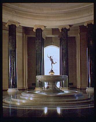 The image shows a grand and ornate room with large columns, marble floors, and elegant architecture. In the center of the space is an impressive fountain featuring Mercury, who stands on top of a shell-like basin as if he's playing his caduceus (staff). The lighting casts dramatic shadows around the statue and highlights its intricate details. This appears to be part of the National Gallery of Art in Washington D.C., specifically depicting Horydczak by approximately 1920-1950, with a focus on Mercury Fountain within that timeframe.