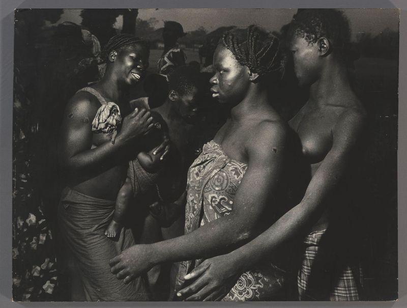 The image is a black and white photograph depicting several individuals, presumably from an African community based on the visual cues such as hairstyles and clothing. There are four people prominently featured in the foreground: two women with their arms around each other's shoulders, standing close together; one woman holding a young child, who appears to be reaching out or moving away slightly from her grasp; and another individual partially visible beside them.

The expressions on the faces of these individuals range from smiles to neutral looks. The clothing style suggests a cultural context, with traditional patterns and fabrics in their attire. In the background, there are other people present but they are less focused and details about them are not as clear due to the depth of field used by the photographer.

The lighting appears natural, indicating that this photo was taken outdoors during daylight hours. The focus is on the interaction among these individuals rather than any specific activity or event. Overall, it captures a candid moment in what seems like everyday life within this community.