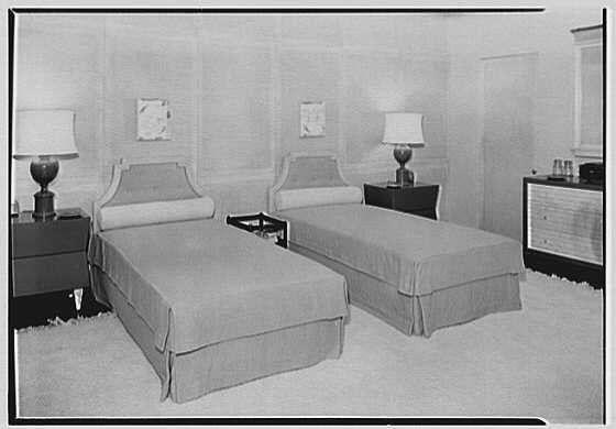 The image shows a room with two single beds placed side by side, each covered in gray bedding. Between the beds are nightstands with lamps on them and what appears to be trays or small tables attached at their headboards. The walls of the room have pictures hanging above the bedposts, and there is a dresser or cabinet against one wall with objects on top. The flooring seems to be carpeted. This black-and-white photograph suggests it may depict an early 20th-century bedroom setting, possibly within a commercial establishment such as a hotel or department store in New York City during April of 1941.
