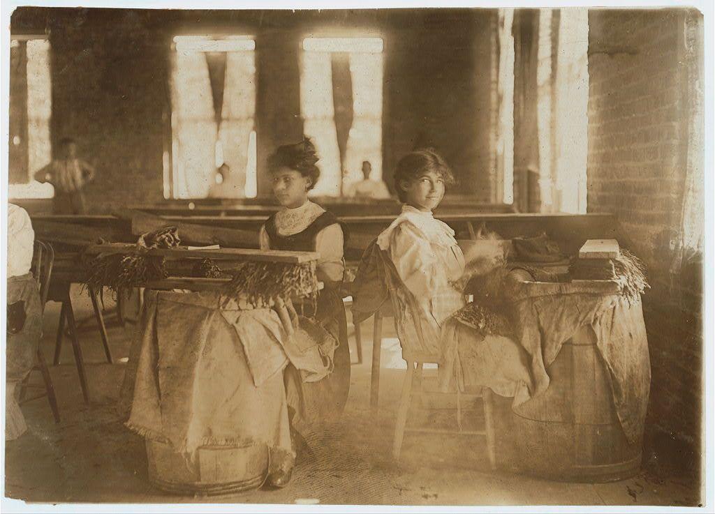 The image depicts two individuals seated at workbenches inside a factory or workshop setting, indicative of early 20th-century industrial practices. The sepia-toned photograph suggests it is historical and possibly documents working conditions during that era.

Both subjects are dressed in period-appropriate attire; the person on the left wears a dark dress with an apron over it, while the individual to the right dons lighter clothing with what appears to be a head covering or bonnet. They seem engaged in tasks involving textiles or fibrous materials, evidenced by their hands working and objects spread before them.

The workbenches are rudimentary, constructed of wood with barrels repurposed as tables; one barrel is draped with fabric. The environment shows windows letting in natural light, a brick wall to the right, and other workers faintly visible through openings or doorways in the background, hinting at an expansive workspace.

Details like shoes on the floor suggest practical working conditions without formal seating arrangements for employees during this period. There's no clear indication of brand names but it portrays a scene that might be part of Lewis Hine’s photographic work documenting child labor and social issues around 1908-1925, focusing on industrial America.

Overall, the image captures a snapshot of early working-class life in an industrial environment [...]