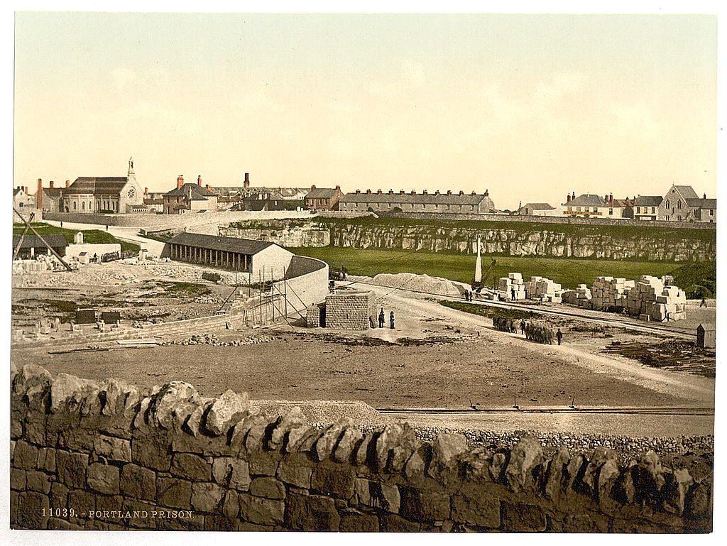The image depicts a historical view of what appears to be an industrial site, possibly from the late 19th or early 20th century. The landscape features open areas with scattered stone blocks and piles, suggesting construction work is underway. A series of buildings in various states of completion occupies the central part of the scene, including what looks like a church or large public building with prominent architecture at the forefront.

In the foreground, a dry-stone wall borders off the area from an elevated position overlooking the site. The background reveals more housing structures and industrial facilities extending into the distance under a clear sky. No people are visible in this snapshot of Portland Prison as mentioned in context provided by caption data but it does show human activity through workers present at the work areas.

This photograph is part of a collection highlighting historical places, specifically from England around the turn of the 20th century and can be found online with specific details pointing to its origins.
