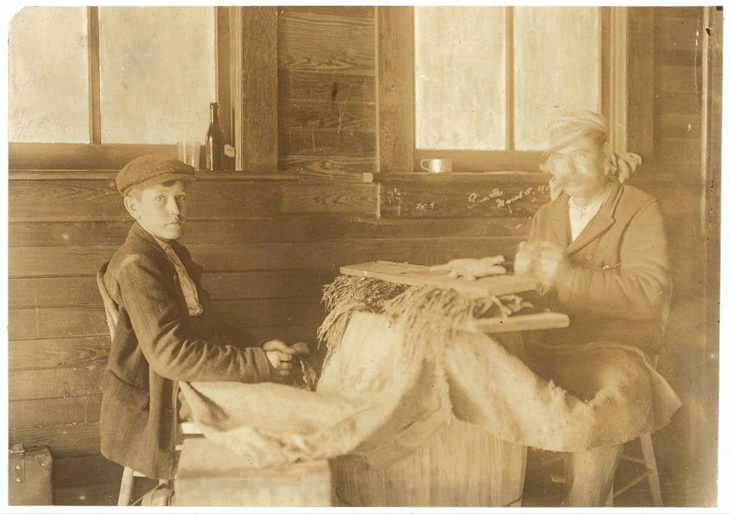 The image is a vintage sepia-toned photograph depicting two individuals seated at what appears to be an indoor wooden table. The person on the left, possibly younger based on their stature and appearance, wears a flat cap, dark coat with visible wear and tear, and trousers tucked into boots. They are holding onto some loose material that drapes over the side of the table, perhaps engaged in a task like mending or sorting textiles.

The individual on the right seems older, wearing a jacket similar to the younger person but without the flat cap, revealing hair pulled back from their face with what could be an apron tied around their waist. They have both hands clasped together and are seated behind more fabric draped across the table in front of them, suggesting they might also be involved in some form of textile work.

The background features wooden-paneled walls, a small window on the far wall showing no visible outside view due to its size or positioning, and an object resembling a bottle placed at a height above eye level. On another surface beside this individual is what appears to be a mug or cup with a handle. There's also part of a suitcase-like item in the lower left corner.

The overall atmosphere suggests a humble domestic setting from perhaps early 20th-century America, where textiles were likely handcrafted items reflecting daily life and labor during that period.