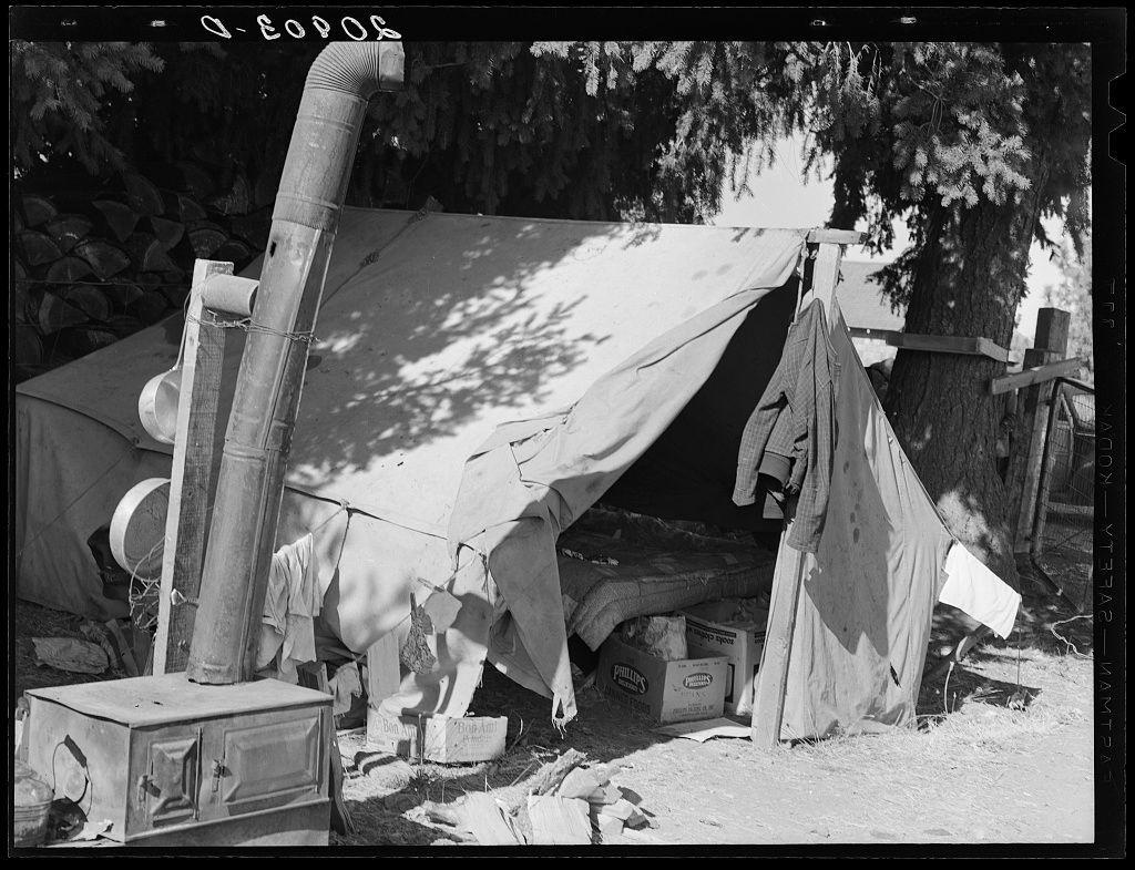 The image depicts a makeshift living space with various objects suggesting habitation. A large, tattered tent is the central focus of the scene, surrounded by assorted items like an old-fashioned stove or furnace, clothes hanging to dry on lines, and wooden pallets leaning against nearby structures. The black-and-white photograph evokes a sense of hardship and simplicity in living conditions during what appears to be challenging times for agricultural laborers, potentially due to economic downturns such as the Great Depression.