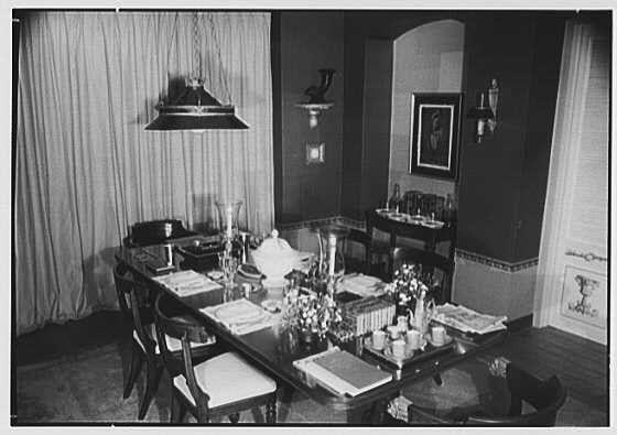 The image depicts a black and white photograph of an elegantly set dining table in what appears to be a vintage home. The room features dark wood paneling on the walls, with decorative moldings at the base and top edges, giving it a classic feel. A large window draped with heavy curtains allows natural light into the space.

The central focus is the long rectangular dining table covered with a white cloth, upon which multiple place settings are neatly arranged. Each setting includes plates, silverware, glasses, folded napkins, and what looks like menus or placemats. The center of the table holds an array of decorative items such as vases filled with flowers, candlesticks with lit candles, books stacked in a pyramid shape, and various other knick-knacks.

In addition to the dining area, there is another smaller round side table next to it that also has more glassware. The overall ambiance suggests a formal or semi-formal gathering could be taking place or about to take place at this time period's aesthetic standards.