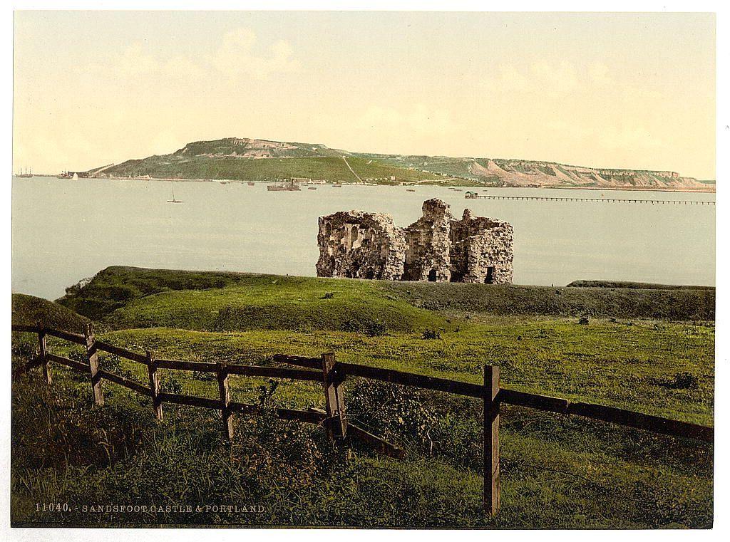 The image depicts a scenic coastal landscape, likely from an older time period given the style of photography and coloration. In the foreground is a wooden fence running horizontally across the bottom third of the photo. Beyond this fence lies expansive greenery that slopes gently towards the water's edge. A large body of calm sea stretches out to meet another landmass in the distance, on which several structures are visible.

Dominating the central area of the image is an old ruin composed of stone walls and remnants of what was likely once a fortified structure or castle. The ruins stand prominently against the green backdrop. In the far background, more hills rise up alongside cliffs adorned with traces of vegetation. A faint outline suggests human activity on these higher grounds.

A small wooden pier extends into the sea from this rocky shore in the distance, and there are several ships visible at various distances within the bay or harbor area beyond it. The sky is largely clear but has a light haze over most of its expanse. There's a sense of tranquility conveyed by the still water and gentle slopes of land.

The coloration suggests that this image might be vintage; earthy greens, browns, and muted blues dominate the scene, giving it an aged look typical of early photographic processes used for such landscapes.