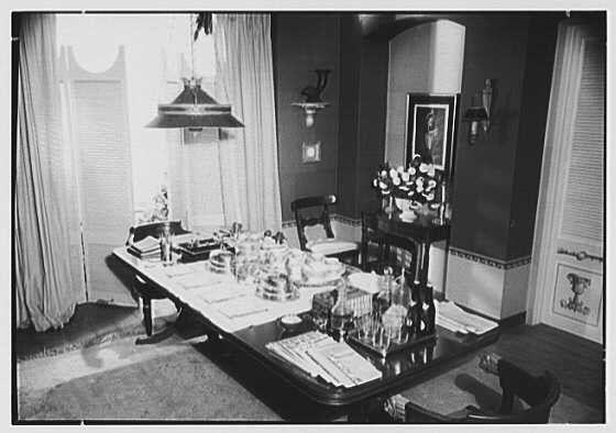 The image shows a vintage black and white photograph of an elegantly set dining table in what appears to be a well-decorated room. The room has large windows with curtains, allowing sunlight to filter through, creating shadows on the walls and floor. A light fixture hangs from one corner of the ceiling.

The dining table is adorned with several items including stacks of plates, silverware, glasses, cutlery wrapped in napkins, a centerpiece arrangement of flowers, and various decorative objects like books and vases. The placement suggests preparation for an event or formal dinner gathering.

Behind the table, there's shelving displaying more glassware and possibly crystal ware. A framed portrait hangs on one wall, adding to the room’s sophisticated ambiance. Other furniture is visible in the background including a chair with plush upholstery by another window.

The overall setting indicates a scene from mid-20th century America, likely 1944 as noted in the caption which also mentions Lord & Taylor's business at Fifth Avenue and Thirty-eighth Street in New York City, suggesting this image could be part of an archive or historical documentation.