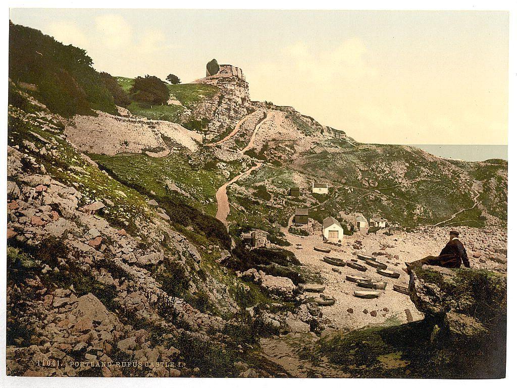This image depicts a picturesque, mountainous landscape with historical significance. It appears to be an old postcard or photograph showcasing Portland Rufus Castle in England. The ruins of the castle are prominently situated on top of a rocky hillside overlooking the sea.

In the foreground, there is a figure sitting on a rock ledge, offering a view down into what seems like a secluded area with small wooden boats lined up along a shoreline. This suggests that this location could be associated with fishing or maritime activities.

The surrounding terrain features rugged cliffs and uneven ground covered in grasses and scattered rocks. A winding path leads to the castle ruins, indicating access for visitors exploring historical sites. The overall atmosphere of the scene is serene yet evocative of past eras when castles played a vital role as fortifications along coastlines.