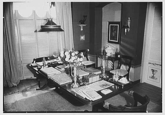 The image depicts an indoor setting, presumably a dining room or study area with antique-style furnishings and decor. A long wooden table occupies the central portion of the frame, upon which various items are arranged including books, papers, vases filled with flowers, glasses, bottles, and what appears to be a teapot set. The scene is well-lit by overhead lamps hanging from a ceiling fixture, casting shadows on the walls behind it.
The room has large windows draped with long curtains allowing natural light to enter through them, reflecting off of the shiny surfaces within the space such as glass objects or polished wooden elements. A portrait hangs on one wall above and to the right side of the table, partially obscured by a standing lamp. The overall color scheme is monochromatic due to the black-and-white nature of this historical photograph.
The image also contains textual annotations that provide context: "Lord & Taylor, business at 5th Ave. and 38th St., New York City," suggesting an association with the well-known retail establishment; "Table set-up III" which indicates a series or documentation purpose; followed by a date stamp of "1944 Aug. 28." Lastly, there is mention of Gottschol-Schleisner Inc., likely referring to the photographer's studio.
The negative dimensions are also provided as safety film with a size indication: "5x7 in."
The source link and image I [...]