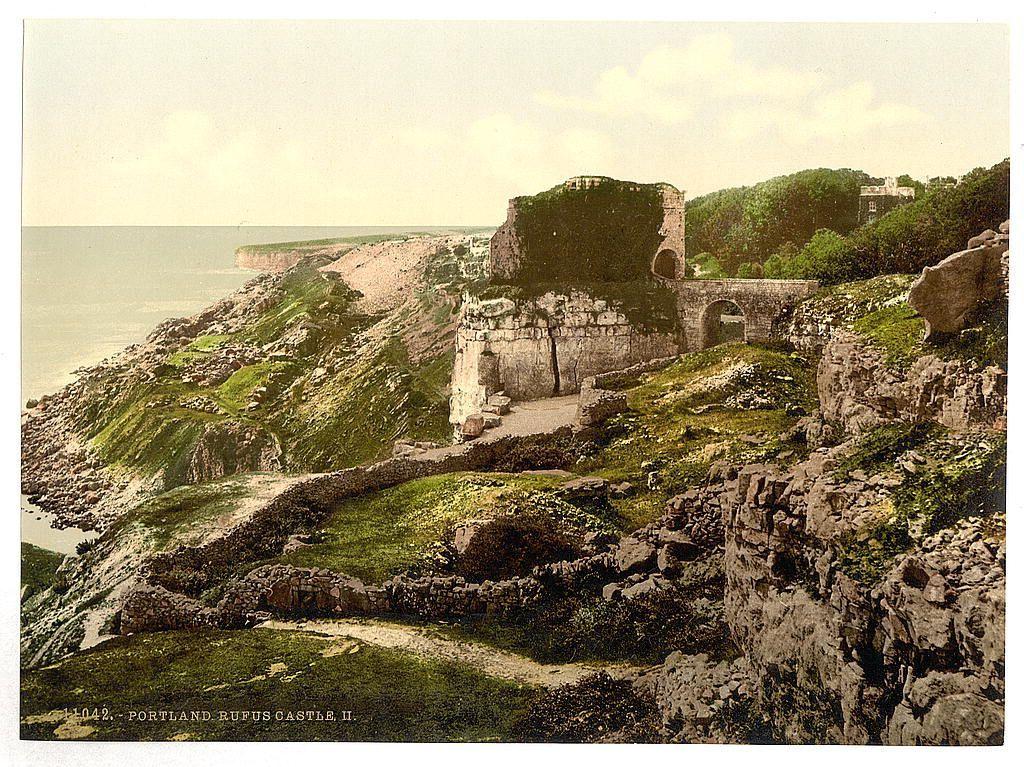 The image displays a picturesque, historic ruin set amidst an expansive natural landscape. It appears to be the remains of Portland Rufus Castle II in England. The structure is composed mainly of weathered stone walls and arches that suggest former grandeur but now stand as fragments against nature's reclaiming forces.

Vegetation has grown on parts of the ruins, indicating their age and exposure to elements over decades or centuries. A pathway winds through the ruins, leading towards a larger building in the distance, hinting at what once may have been its purpose — perhaps residential or administrative.

The backdrop is dominated by rolling hills covered with greenery and trees, which contrast against the clear sky above. The coastline can be faintly seen on the horizon line where the land meets the sea, adding to the castle's isolated yet commanding presence over the landscape.