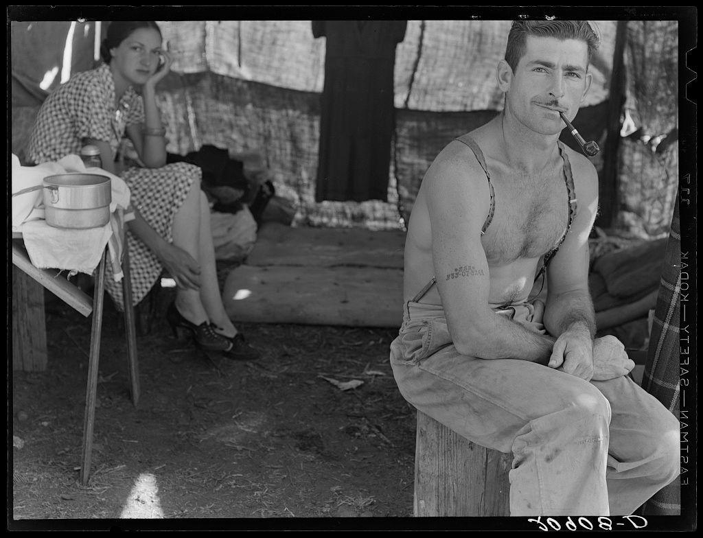 The image is a black and white photograph depicting two individuals, presumably an unemployed lumber worker and his wife, as they go to the bean harvest. The man sits on a makeshift stool outside what appears to be their humble dwelling with curtains made of fabric or paper over wooden supports behind him. He has a rugged appearance, shirtless except for suspenders holding up his trousers, revealing visible tattoos including one spelling out "EVE" across his right shoulder and arm. A notable feature is the social security number tattooed on his left forearm.

He holds what seems to be a pipe in his mouth, suggesting he may have been smoking or plans to smoke it soon. His expression is neutral but perhaps contemplative, indicative of the hardships they might face as unemployed workers during challenging economic times like those seen during the Great Depression era.

The woman sits on a chair at a table covered with a checked cloth beside him, draped over her shoulder and legs are items that appear to be personal effects or tools. She is dressed in what looks like an apron patterned dress, wearing shoes indicative of modest footwear for working conditions. Her posture shows contemplation as she rests one hand on the side of her face.

In the background, we can see part of their dwelling with a curtain-like fabric covering windows and perhaps providing privacy or shade. The [...]