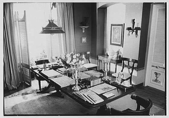 A black and white photograph of a dining room table set up for an event, possibly in 1944 as indicated by the caption. The table is arranged with various items such as books, decorative vases, flowers, glasses, cutlery, and placemats on it. Curtains hang near windows allowing natural light into the room. There are chairs around the dining area which seems to be in a formal setting possibly for an important event or gathering.