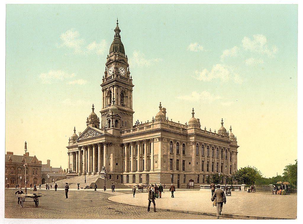 The image shows a historical photograph depicting a grand, ornate building with multiple columns and domed structures. Prominently featured is an imposing clock tower rising from the center of this structure. The architecture suggests it could be significant for civic or governmental functions. In front of the building is a spacious area paved in what appears to be cobblestones, where people are scattered around: some walking individually while others are gathered near benches and trees. There's also a horse-drawn cart on one side, hinting at transportation methods from an earlier era.

The photograph has a vintage quality with sepia tones dominating the color scheme, suggesting it was taken in or before the 20th century. The clarity of the image hints that it may have been professionally printed using techniques like photochrom printing popular around late 19th to early 20th centuries. This form of printmaking could explain why details such as individual faces and finer textures are not sharply clear.

Given these visual cues, this structure is likely Portsmouth's Town Hall in the UK. The presence of people dressed in period attire further supports its historical context.