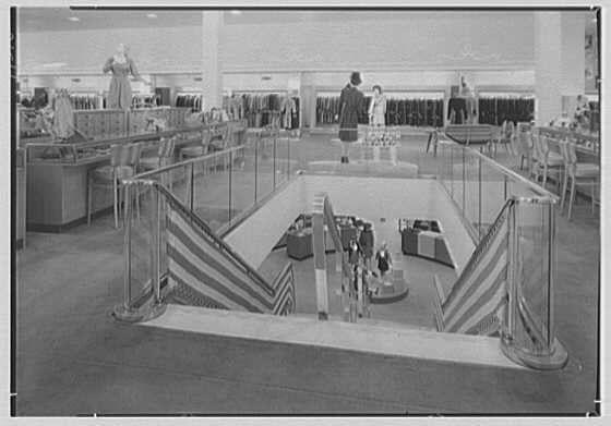 The image is a black and white photograph depicting the interior of a large department store. The scene shows an escalator with striped pattern railings leading to different levels within the building, suggesting it's designed for pedestrian traffic between floors. Several people are visible in various areas throughout the space: some appear to be shopping or browsing items displayed on racks along one wall, while others seem engaged in conversation near a counter.

In addition to the escalator and shoppers, there is an information desk with several individuals gathered around it, possibly assisting customers with inquiries or directions. The store's layout includes multiple aisles filled with merchandise, which are organized into categories for easy navigation by visitors.

Overall, this image provides insight into the retail environment of a mid-20th century department store and how people interacted within such spaces during that era.