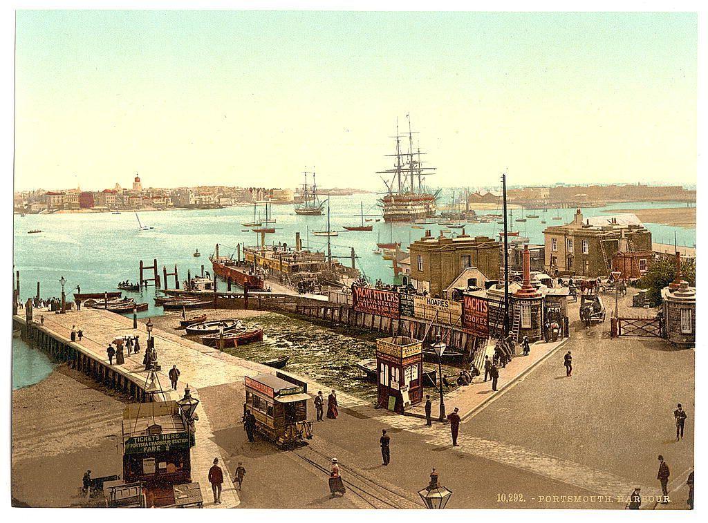 The image depicts a bustling harbor scene in what appears to be early 20th-century England. In the foreground, there is a busy street lined with various establishments such as "Tickets Here," which suggests ticket booths for travel services or possibly theater entrances, and other signs indicating shops selling items like yachts and hooks. People are walking along the streets and sidewalks, some pushing handcarts.

The harbor itself is active with numerous boats docked at wooden quays, including a prominent sailing ship in its midst. Larger ships can be seen anchored further out to sea under an overcast sky. In the distance across calm blue waters lies more of Portsmouth's coastline featuring buildings with distinctive architecture typical for that era and region.

The coloration is faded but suggests sepia tones common to old photographs, indicating this might not have been colored when it was taken or has aged in such a manner. The image carries historical significance as part of the Photochrom prints series from around 1890-1900, which were known for their detailed and vibrant color reproductions based on photographic negatives.

The photograph is attributed to Loener, with reference details indicating it's located within an online archive or collection dedicated to such images. The overall atmosphere exudes a sense of bygone era life in England’s bustling ports during [...]