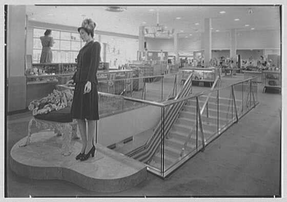 The image depicts a vintage scene from an indoor setting, likely a department store or shopping center. The focus is on two women dressed in elegant attire of the mid-20th century. One woman stands prominently at the forefront, examining a display table laden with what appears to be dolls or small toys. She wears a dark dress and holds onto the railing as she peruses her items.

Behind her, another figure can be seen looking through windows towards an outdoor area where more people are visible in the distance. The overall ambiance suggests a mid-20th-century retail environment characterized by its spacious layout with multiple floors accessible via escalators or staircases equipped with safety railings for customers' convenience and protection.

The image is monochromatic, emphasizing grayscale tones typical of early black-and-white photography from that era. Additional context provided indicates this photograph was taken in August 1946 at Lord & Taylor's business location on Long Island, New York. The visual details evoke a sense of nostalgia for the bygone days of fashion and retail culture during World War II.