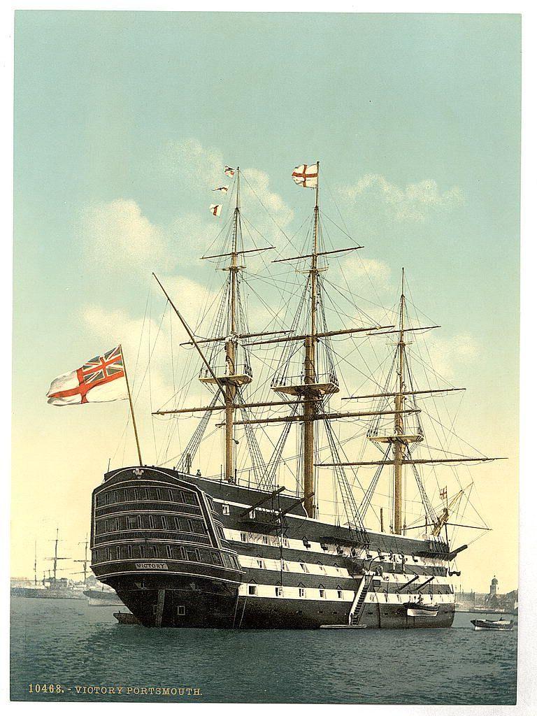 The image depicts a majestic sailing ship with multiple masts and complex rigging, prominently featuring the Union Jack flag on its starboard side. The vessel is anchored in calm waters, suggesting it's moored at port rather than cruising or engaged in active naval maneuvers. It appears to be an old-fashioned warship, possibly from the 18th century given its design. In the background, other ships and small boats are visible along with a distant shoreline that includes buildings indicative of a cityscape. The sky is partly cloudy but predominantly blue, suggesting fair weather conditions at sea during daylight hours. A date "VICTORY PORTSMOUTH" and a number "10468" can be seen on the image indicating its historical context or edition as a print postcard. There's also an indication of it being in Portsmouth England which is known for its historic naval significance, possibly referencing this ship to Victory (ship, 1765), Nelson’s flagship during various campaigns including at Trafalgar.