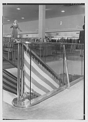 This black and white photograph depicts a woman standing on an escalator within what appears to be a department store or shopping center. The escalator is striped with alternating wide bands of dark color, possibly red and grey, which create the illusion of movement as they descend downwards.
The woman has blonde hair styled in waves typical of mid-20th-century fashion trends. She's wearing a dress that falls above her knees, paired with heels, indicative of women's clothing from around 1946. The escalator is framed by large windows or glass barriers on both sides and appears to be empty except for the woman.
In the background, other store patrons can be seen browsing through garments displayed in what seems like a clothing section. Some racks are visible with hangers displaying various articles of clothes hanging down from them. On one side, there is another escalator going upwards which suggests that this area serves as an intermediate floor within the multi-level building.
The lighting appears to come primarily from overhead sources, illuminating the space evenly and highlighting reflections on the shiny surface of the escalators' steps. The overall atmosphere conveys a sense of modernity and progress for its time period, reflecting 1940s architecture in America with an emphasis on convenience and accessibility through amenities like escalators within retail environmen [...]