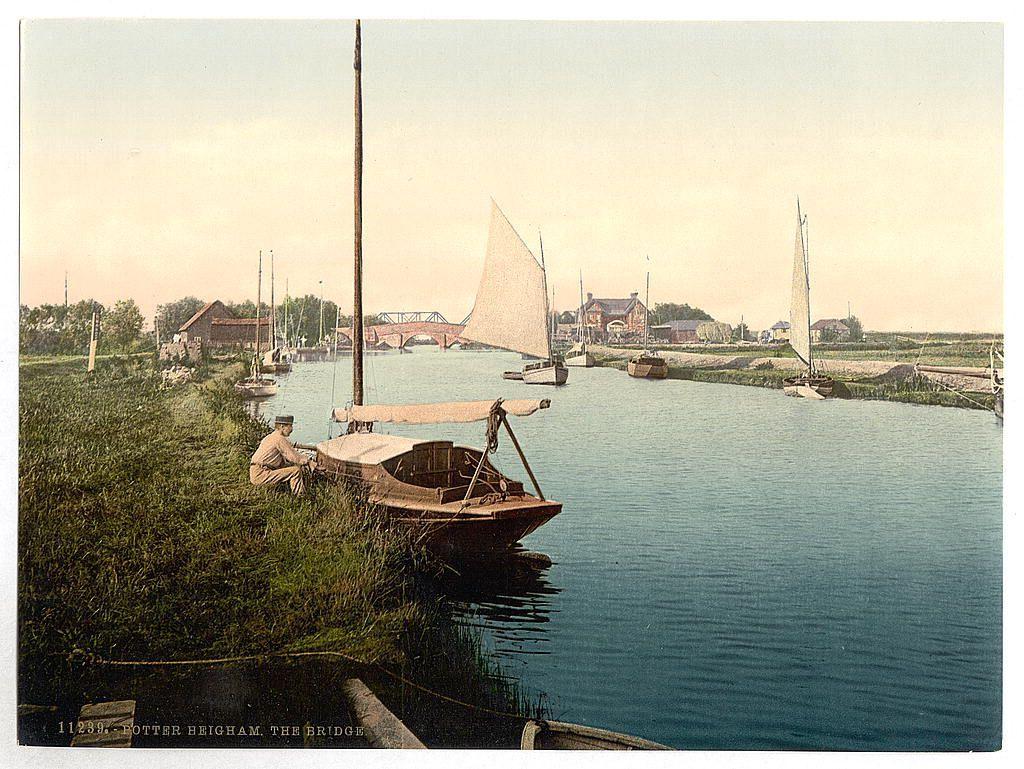 This image depicts a peaceful riverside scene with various boats moored along the water's edge. A man is sitting on one of these boats, possibly engaging in maintenance or simply enjoying the view. The river extends into the distance where bridges and buildings can be seen across it, suggesting this may be a significant local thoroughfare or landmark. The overall atmosphere appears tranquil and leisurely, with no signs of industrial activity or modern infrastructure that would indicate any time later than early to mid-20th century at latest.