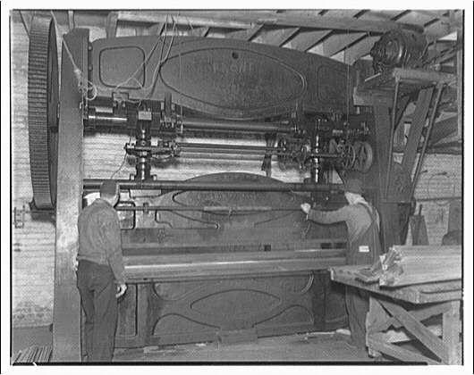 A black and white photograph depicting a vintage industrial scene with two individuals operating heavy machinery, which appears to be part of an assembly line. The machine consists of large metal components arranged vertically in pairs, possibly indicating its use for manufacturing or construction purposes. Each pair is connected by chains or belts that transfer mechanical power from one component to another.
The image shows a man on the left side wearing casual work attire including trousers and a long-sleeved shirt with rolled-up sleeves, standing beside what seems like an elevated platform where his hands are placed near some components of the machine. Another individual is seen on the right interacting with parts of the machinery closer to ground level.
In the background, other industrial equipment can be observed alongside brick walls indicative of older building structures and large wooden beams above suggesting a spacious environment typical for manufacturing or maintenance activities during that era. The scene exudes an atmosphere characteristic of early 20th-century workshops where manual labor played a crucial role in industrial processes.