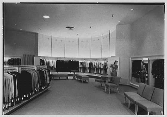 The image depicts a spacious clothing store interior with rows of neatly arranged garments displayed on hangers. The lighting is bright and even, illuminating the various sections within the shop. In one section, there's an array of suits or dress pants hanging in different shades of color along shelves. Another area shows dresses draped over chairs. A few seating arrangements are provided for customers to try on clothes comfortably.

The layout includes open aisles that allow easy movement through the store and access to various products. The overall design is clean, modern, and functional with a focus on organization which seems typical of mid-20th-century retail stores aiming to create an inviting atmosphere for shoppers. No people are visible in this particular shot; only one person can be seen at a distance reflecting herself in the large mirror.

The image carries historical significance as it is part of Lord & Taylor's business documentation, specifically from their Boston Post Rd and Wilmot Rd location in Scarsdale, New York during February 24th, 1948. The photograph was taken by Gottschalk-Schleisner Inc., indicating professional photography services used for documenting or marketing purposes at that time.

The image is a monochromatic snapshot captured on safety film with dimensions of approximately 5x7 inches, showcasing the detailed interior design and merchan [...]