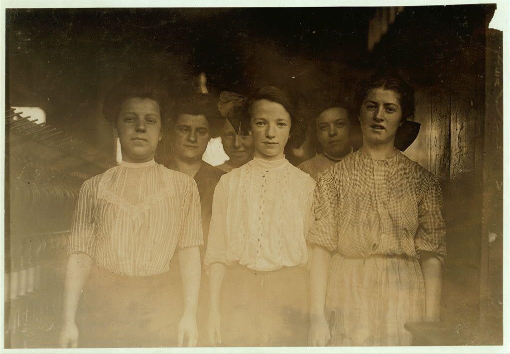 The image is a historical black and white photograph depicting six individuals who appear to be women, likely from the late 19th or early 20th century. They are standing close together in what seems to be an industrial setting with dim lighting, possibly within a factory environment like Brookside Worsted Mills as mentioned in the caption.

The group is dressed in simple work attire suitable for manual labor; some wear striped shirts while others have white blouses and aprons over their clothes, which were common during that period. Their expressions are neutral to slightly serious, suggesting they might be at a moment of rest or pause from working within the mill's textile production.

In terms of context provided by the caption, this photograph captures one of the youngest groups of spinners in Brookside Worsted Mills located in Westford, Massachusetts. The specific focus on their youthful age is emphasized through the mention that they are among "the youngest there," indicating a sense of youth or perhaps highlighting working conditions for younger individuals during that time.

For visually impaired users: This image shows six women standing close together; possibly in an industrial setting from late 19th to early 20th century. They wear simple work attire with stripes and white blouses, indicative of textile workers at Brookside Worsted Mills in Westford, Massachuset [...]