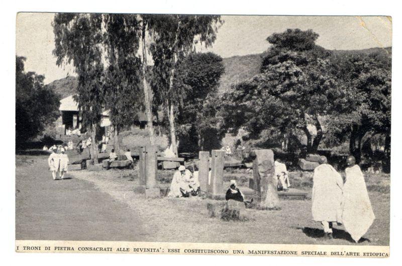 The image displays a black and white photograph of an outdoor setting with several people engaging in various activities. In the foreground, two individuals dressed in traditional Ethiopian robes are walking away from the camera on what appears to be a path or road. Behind them, there is another pair of figures conversing while seated under trees.
In the middle ground, multiple stone structures and crosses suggest that this location could be a cemetery dedicated for sacred purposes. Some people can be seen sitting near these stones, possibly reflecting or mourning. A group sits in contemplation on a bench amongst the grassy area with some standing nearby.

The background showcases lush greenery and trees, hinting at a serene rural environment. The overall atmosphere of the photograph exudes calmness and reverence, indicative of an Ethiopian cultural site dedicated to religious observance.
At the bottom right corner of the image is text in Italian that reads "I TRONI DI PIETRA CONSACRATI ALLE DIVINITA': ESSI COSTITUISCONO UNA MANIFESTAZIONE SPECIALE DEL'ARTE ETIOPICA," which translates to a description about sacred stone altars dedicated to the divinities and their special artistic manifestation in Ethiopian art. This text provides context for understanding that the setting is linked with religious or cultural significance within Ethiopia, showcasing an example of traditio [...]