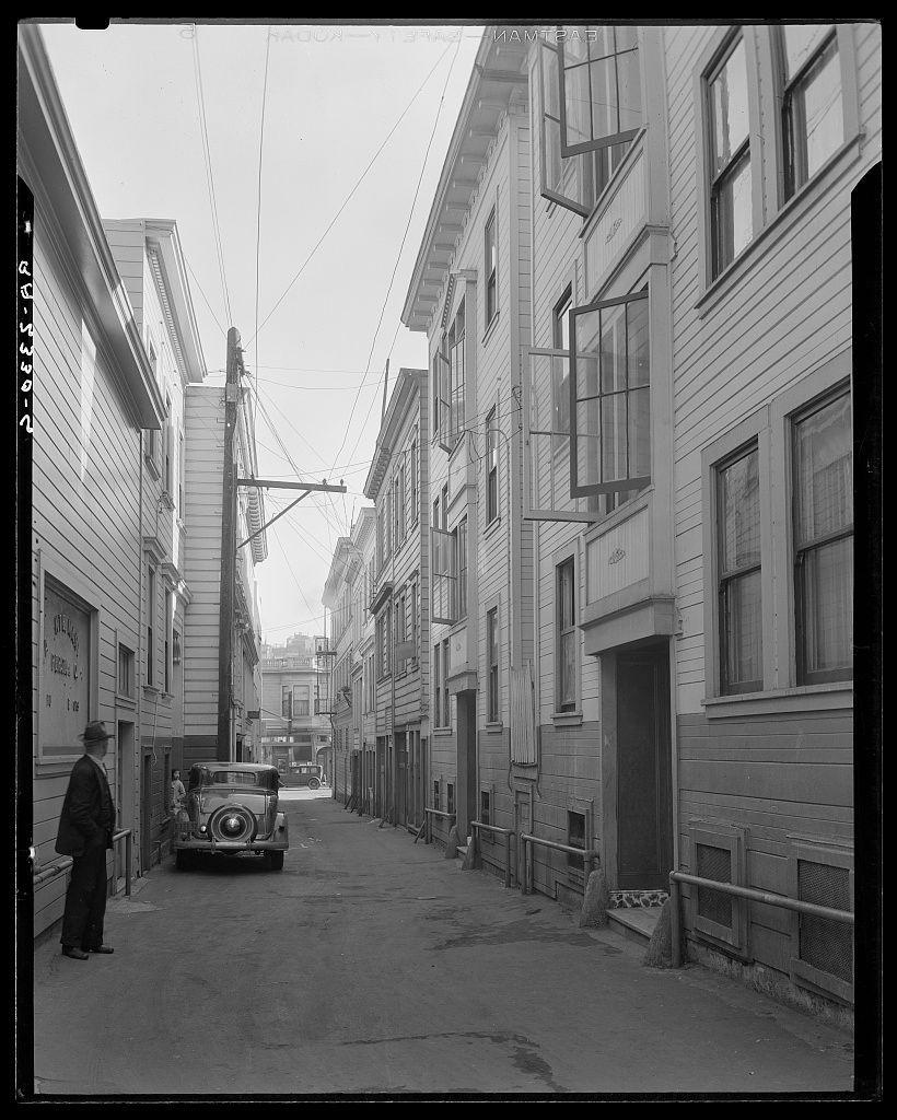The image shows a black and white street scene from an alleyway in San Francisco, California. The architecture is characterized by narrow buildings with wooden facades and metal railings on the sides of some windows. A vintage car parked at the end of the alley catches attention due to its classic design featuring round headlights and chrome accents.

On the left side of the image, a figure appears to be walking down the street, dressed in dark clothing that blends with shadows cast by overhead wires or power lines extending from buildings above. The overall atmosphere is quiet and somewhat desolate, suggesting it could have been taken during an early morning or late afternoon when foot traffic was minimal.

The vintage car's presence suggests this photo might date back several decades ago. Overall, the image captures a moment in time within San Francisco’s Italian district known as North Beach (or "North Bean," possibly indicating historical documentation). The setting reflects urban decay and contrasts with modern-day cityscapes typically associated with bustling streets filled with people.

Additional context for understanding this photo can be found at Card Alley.