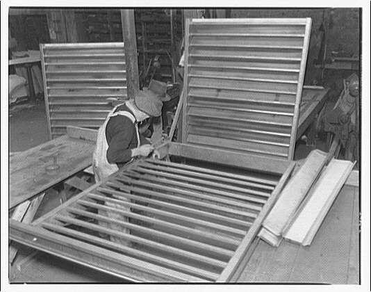 The image shows a black and white photograph of an individual working on the construction or assembly of large metal ventilators in what appears to be an industrial workshop setting. The person is wearing protective gear, including gloves, overalls with suspenders, and possibly safety goggles around their neck. They are using tools to work meticulously on one of several panels that form part of the vent structure.

In the background, other similar vents or parts can be seen placed in various orientations, indicating an organized assembly area likely within a factory environment dedicated to producing such equipment. The overall atmosphere suggests manual labor and craftsmanship from what seems like early 20th-century industrial manufacturing practices.