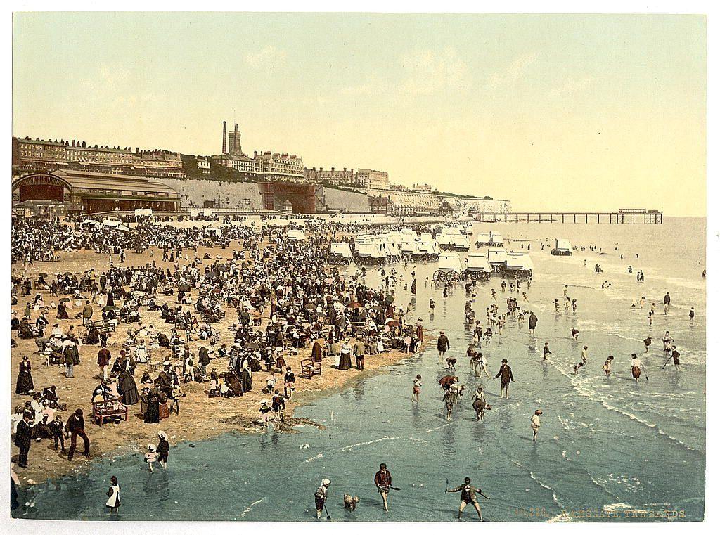 A vintage postcard or photograph depicting a bustling beach scene with numerous individuals and groups scattered across the sandy shore. The image shows people engaging in various activities, such as walking along the water's edge, sitting on chairs, playing games, sunbathing, and socializing. Some are dressed in period attire suggesting an early 20th-century setting.

The backdrop features a pier extending into the sea with figures likely enjoying rides or strolling around. Behind this scene is a row of buildings that could be part of beachside establishments like hotels or amusement venues. A prominent structure stands out, possibly a tower or landmark associated with Ramsgate in England during the late 19th century to early 20th-century period.

The color palette includes muted sepia tones typical of older photographs, giving it an antique feel. The presence of horse-drawn carriages and other vintage elements adds authenticity to this historic snapshot capturing a day at the seaside in Ramsgate, England around the turn of the previous century.