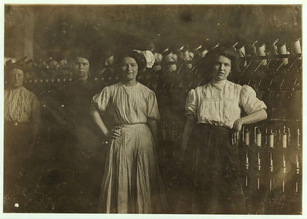 The image depicts a group of women working in what appears to be an industrial setting, likely a textile mill based on the spindles and reels visible in the background. The photo is black and white, suggesting it was taken during the late 19th or early 20th century when such mills were common places for labor-intensive work.
The women are dressed in modest, utilitarian clothing typical of working-class attire from that era; they wear long-sleeved blouses with gathered sleeves at the wrists and waist-length skirts. Their expressions range from neutral to mildly concerned, suggesting a sense of seriousness or perhaps fatigue related to their work. The lighting is dim, casting soft shadows on their faces, which adds an air of solemnity to the scene.
The background features rows upon rows of spindles with white coils wound around them, indicating that this was likely a yarn-spinning operation within a larger textile mill complex. These mills were critical in the industrial revolution for producing textiles such as woolen goods.