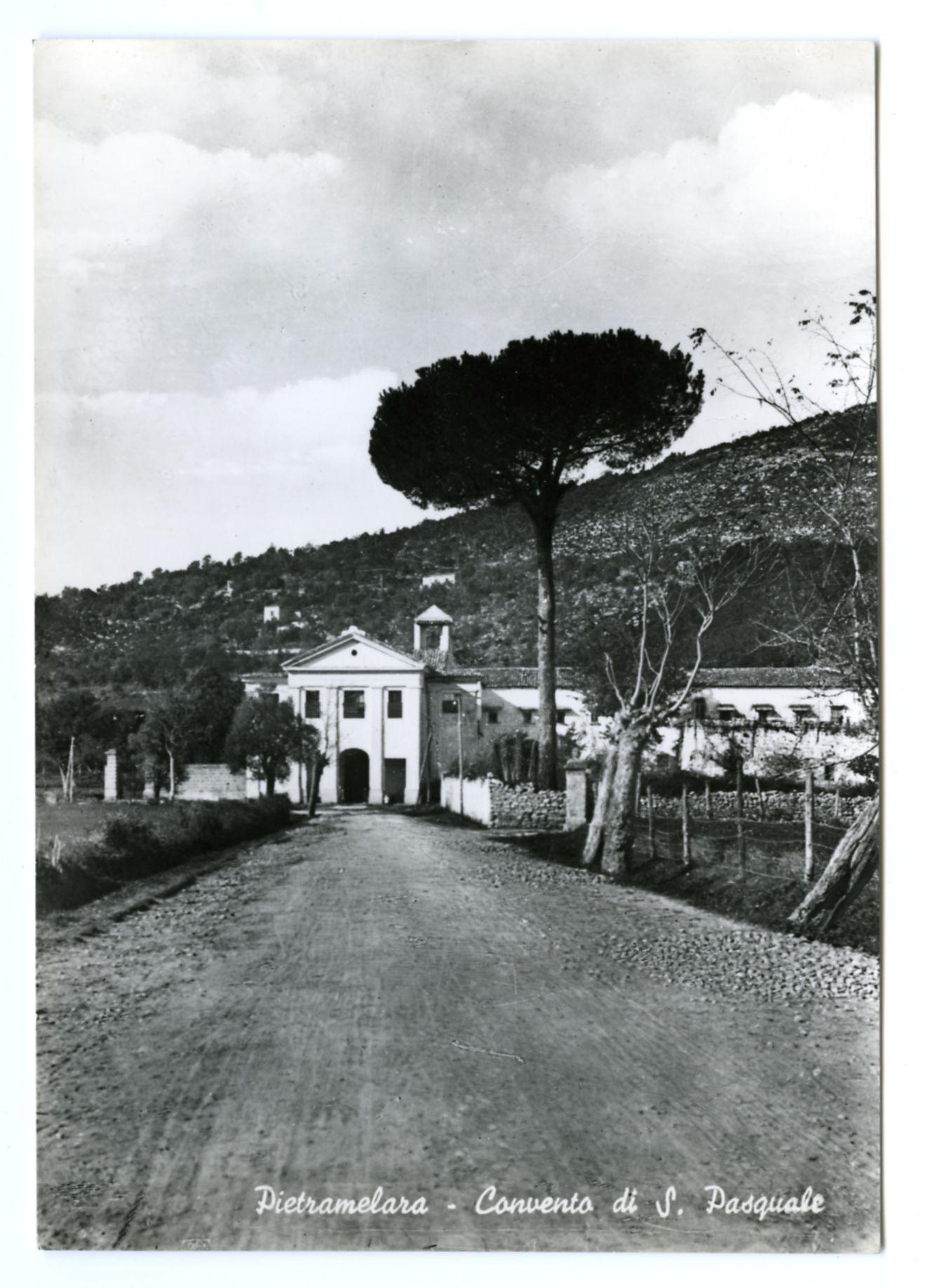 The image depicts a black and white photograph of an old, rural landscape with a historic building in the center. The structure appears to be a convent or monastery named "Convento di S. Pasquale," as indicated by the text at the bottom right corner of the photo. A prominent evergreen tree stands on one side of the road leading up to the convent, and other trees are scattered around the area.

The building itself has two main sections: a central part with what seems like arched entryways or windows, possibly made from white stone or plaster, and an adjacent structure that appears more utilitarian. The architecture suggests it may be several decades old, with classical influences such as columns on either side of one entrance. A bell tower is visible in the background atop another building.

The surrounding area has a hilly terrain covered with vegetation, typical of a countryside setting or semi-mountainous region. There's also some fencing and what appears to be agricultural fields at various distances from the convent. The sky overhead shows clouds indicative of an overcast day.