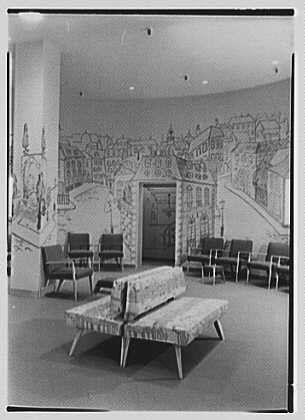 The image is a black and white photograph depicting an interior space, likely within a business or public building from the mid-20th century. The setting features a large mural on one wall that illustrates what appears to be an urban landscape with various buildings, pathways, and people engaged in different activities.

In front of this mural sits a bench-like furniture arrangement made up of several individual chairs arranged around a central square table. These pieces are covered with patterned fabric, possibly indicating they serve as waiting or lounge seating for visitors.

The floor is polished concrete, reflecting the overhead lights that illuminate the space from recessed fixtures in the ceiling. The overall ambiance suggests an environment designed to be comfortable and aesthetically pleasing for guests or clients of a particular establishment.

Additional information indicates this photograph was taken at Lord & Taylor's business located on Boston Post Road and Wilmot Road in Scarsdale, New York specifically within their shoe department on the main floor during February 24th,1948. The image is credited to Gottschol-Schleisner Inc., a company known for producing architectural photography.

Based on these details, it can be inferred that this photograph captures an elegant and thoughtfully designed space meant to enhance customer experience within Lord & Taylor's  [...]